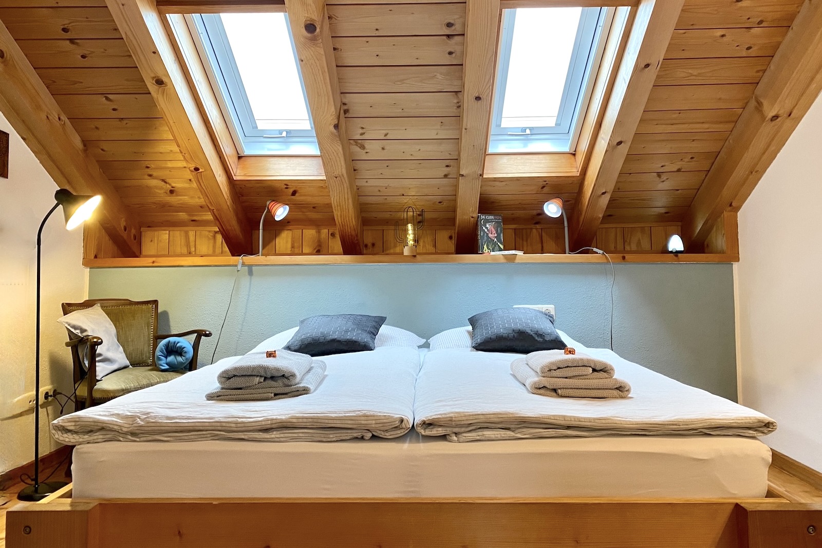 Schlafzimmer mit Doppelbett unter Holzdachstuhl mit Dachfenstern vor blauer Wand.